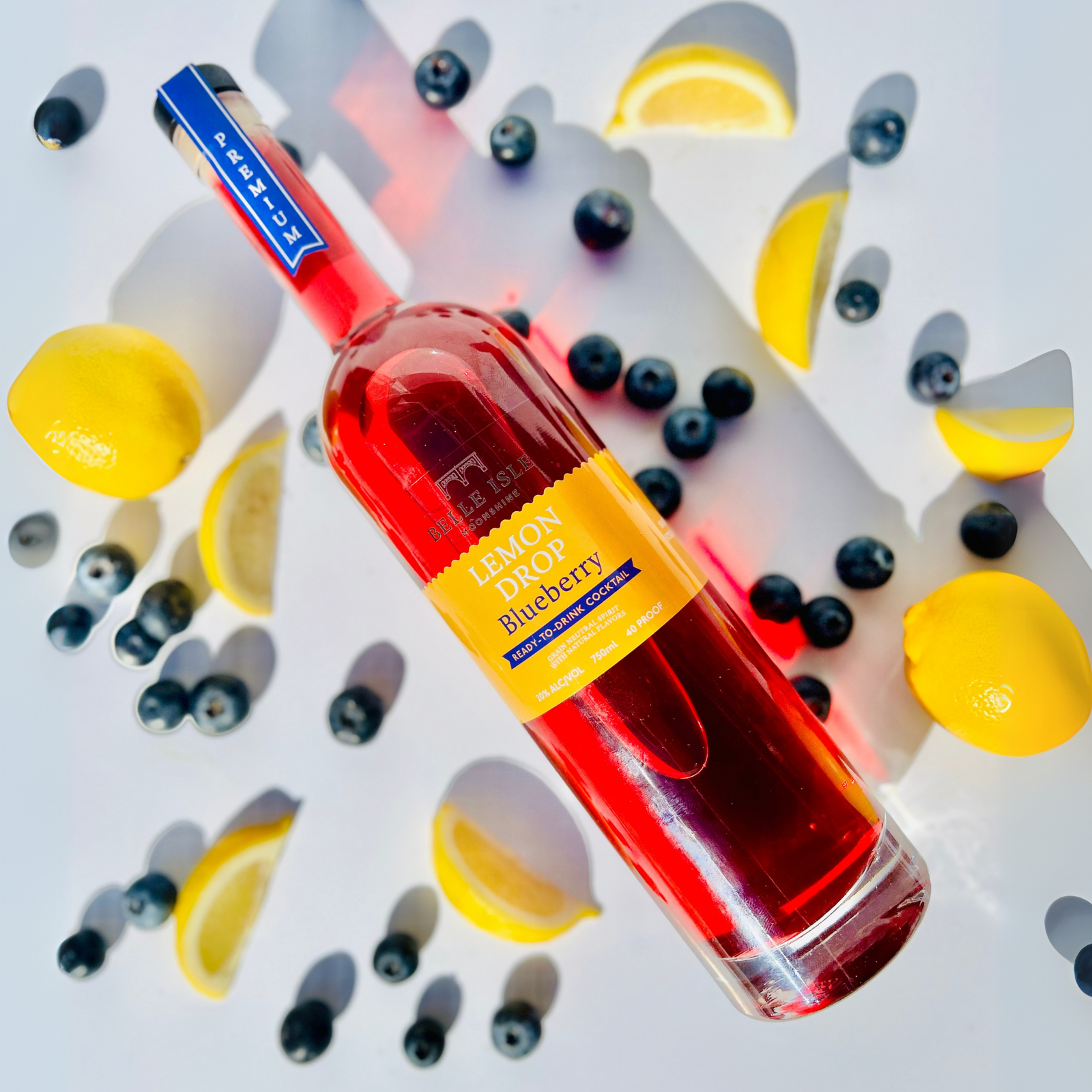 A bottle of Belle Isle Lemon Drop Blueberry moonshine with a backdrop featuring blueberries and lemon slices. The bottle is prominently in the center with a red tinted liquid visible through the clear glass.