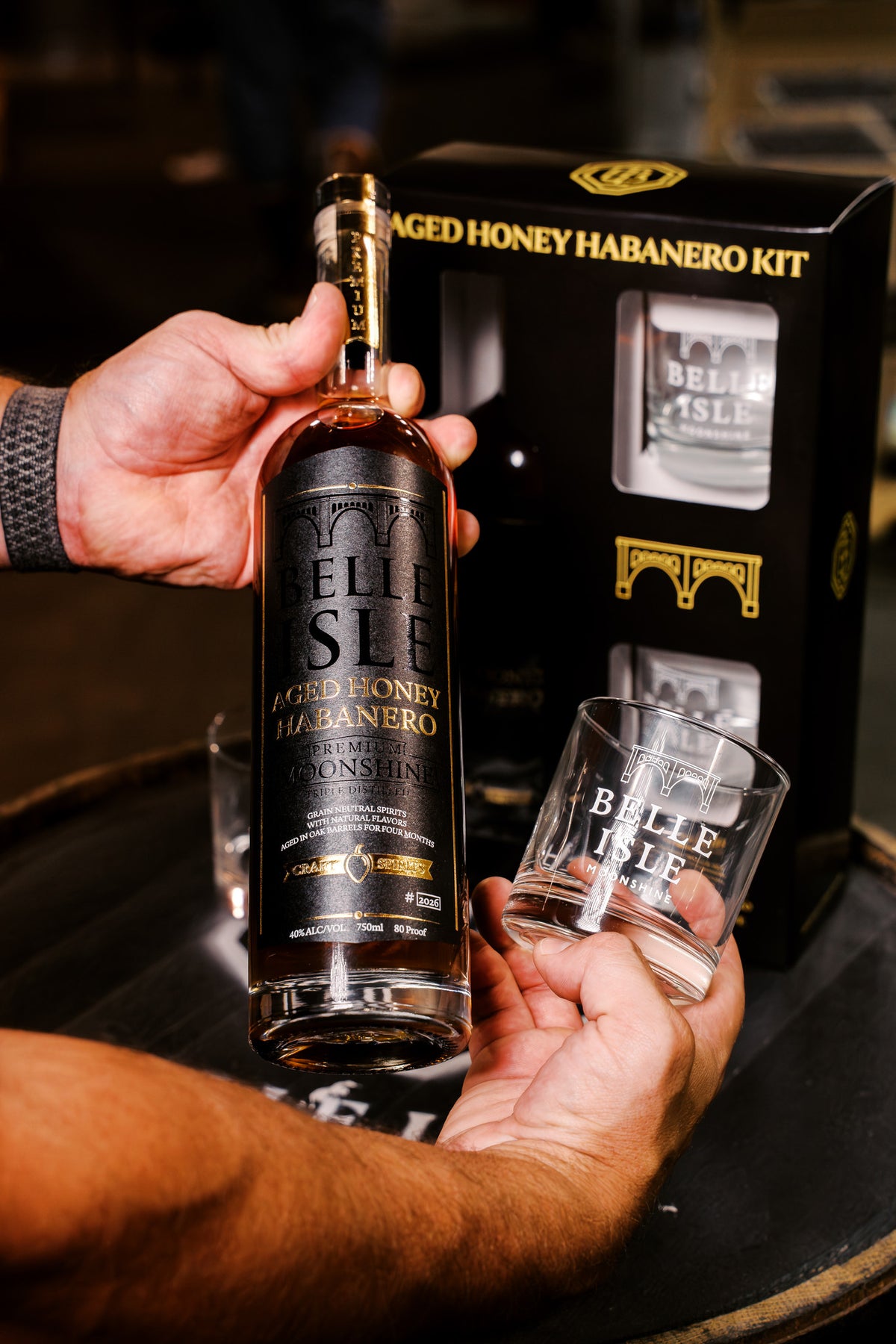 Hands holding a bottle of Belle Isle Aged Honey Habanero and an etched Belle Isle Moonshine rocks glass in front of the kit box